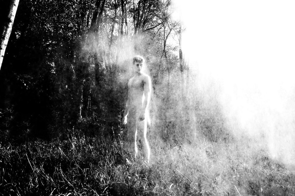 A black and white photograph of a nude young man fading into the mist where a grassy field meets a forest.